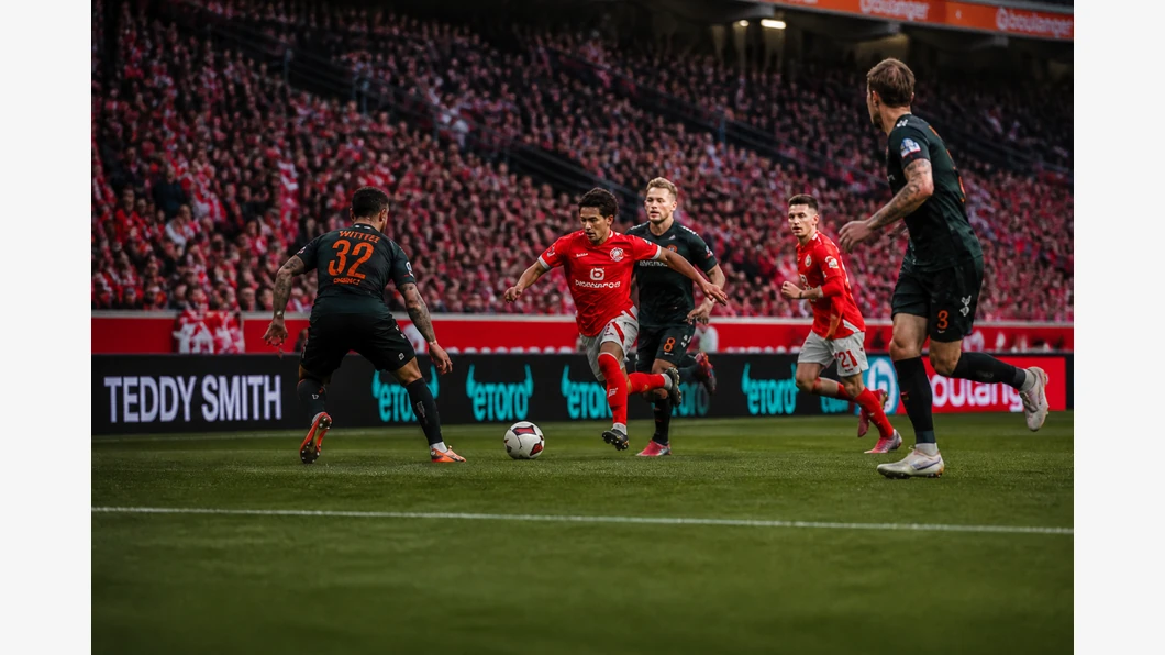 Anthony Caci de Mainz et Marco Grüll de Werder se disputent le ballon au premier plan, devant des tribunes floutées.